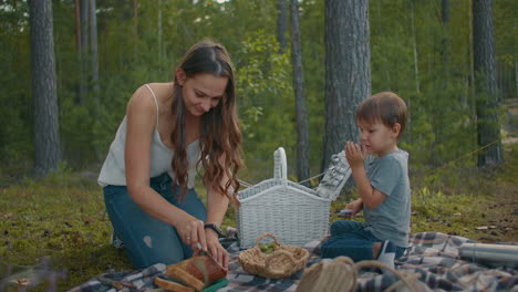 young-woman-is-cutting-bread-in-picnic-little-boy-is-sitting-near-on-blanket-family-rest-in-forest-at-summer